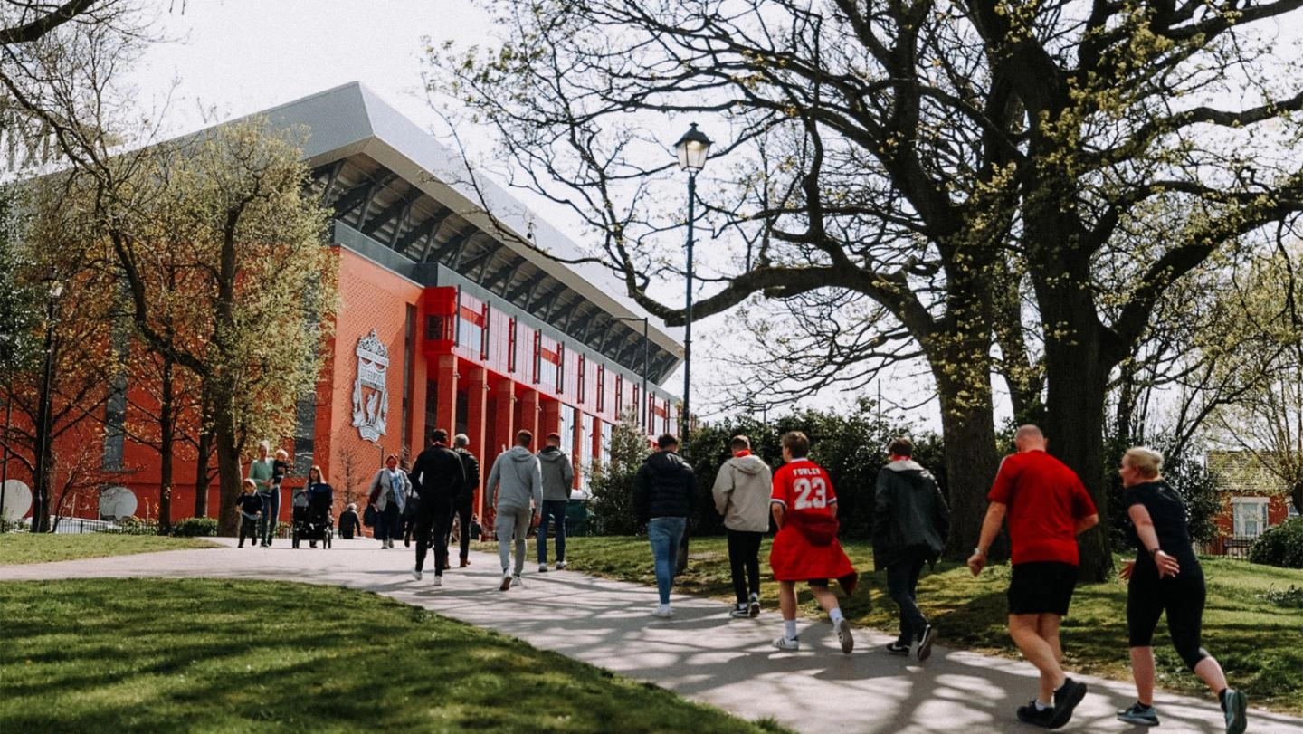 People walking up a path in a park surrounded by trees, towards Anfield Stadium