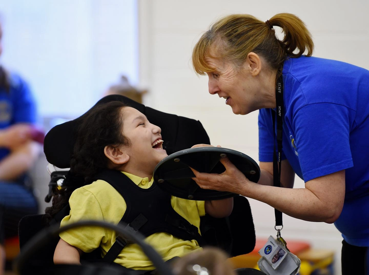 A woman in a blue shirt interacts with a smiling child in a wheelchair